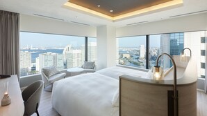 A modern hotel room with a large bed, a desk, a chair, and a view of the cityscape.