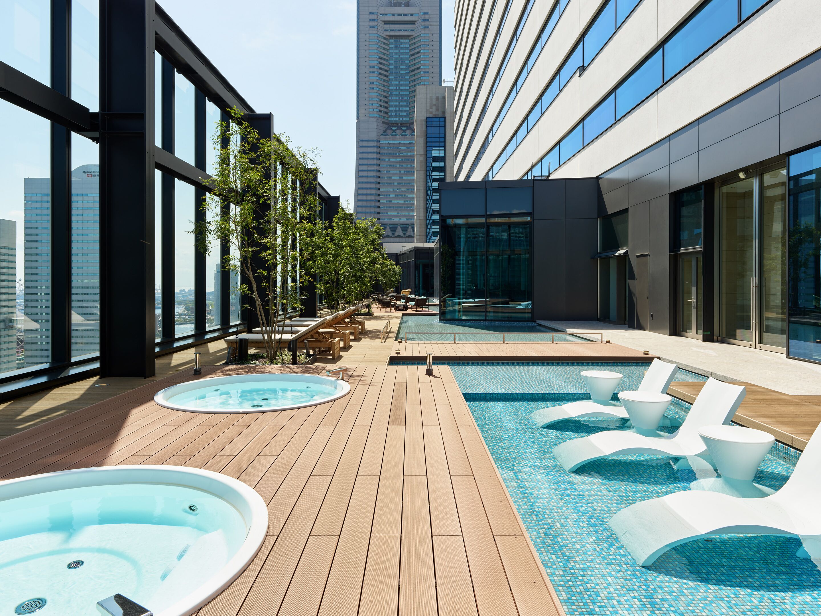 A rooftop pool area with lounge chairs and a hot tub, surrounded by large windows offering city views.