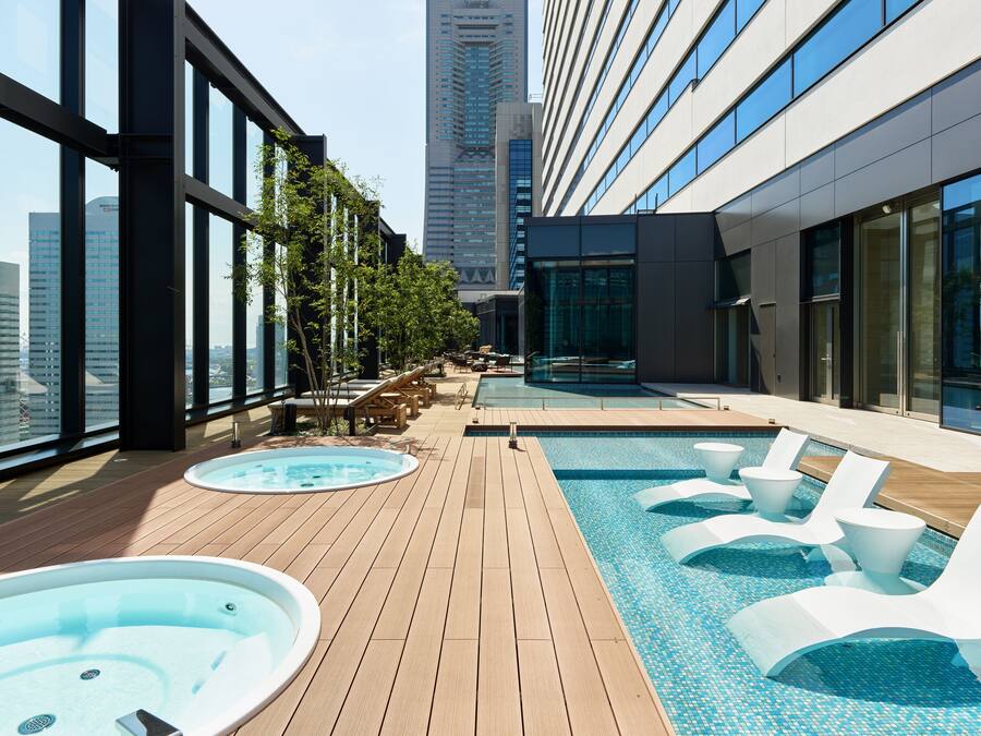 A rooftop pool area with lounge chairs and a hot tub, surrounded by large windows offering city views.
