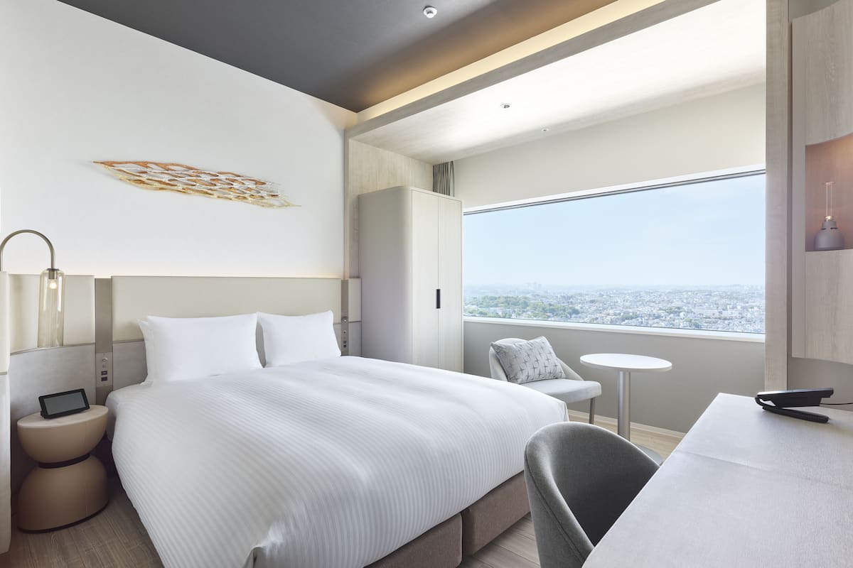 A modern hotel room with a large bed, a desk, a chair, a nightstand, and a view of the city.