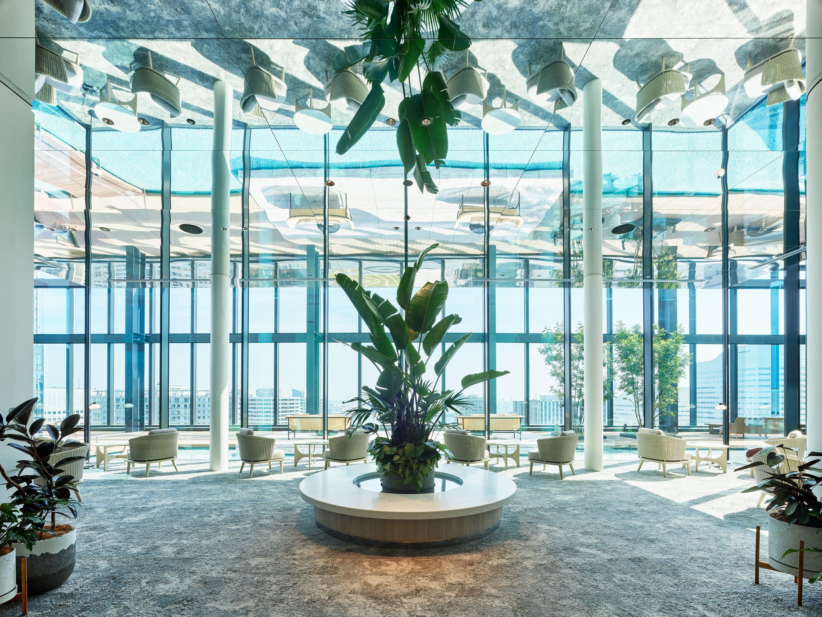 Un espace intérieur vaste et lumineux, doté d’une grande fenêtre, d’un espace de sièges circulaire central et de plusieurs plantes en pot.