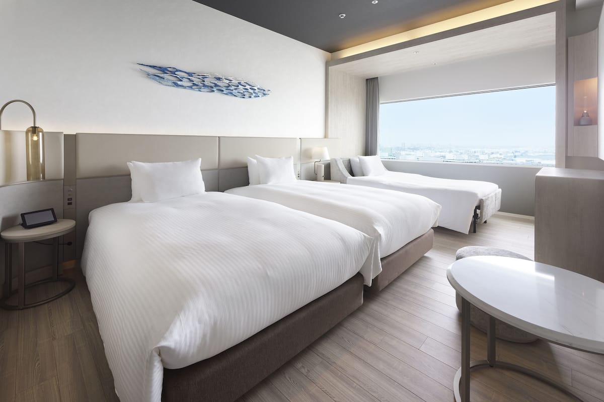 A modern hotel room with two beds, a small desk, a tablet, and a view of the city.