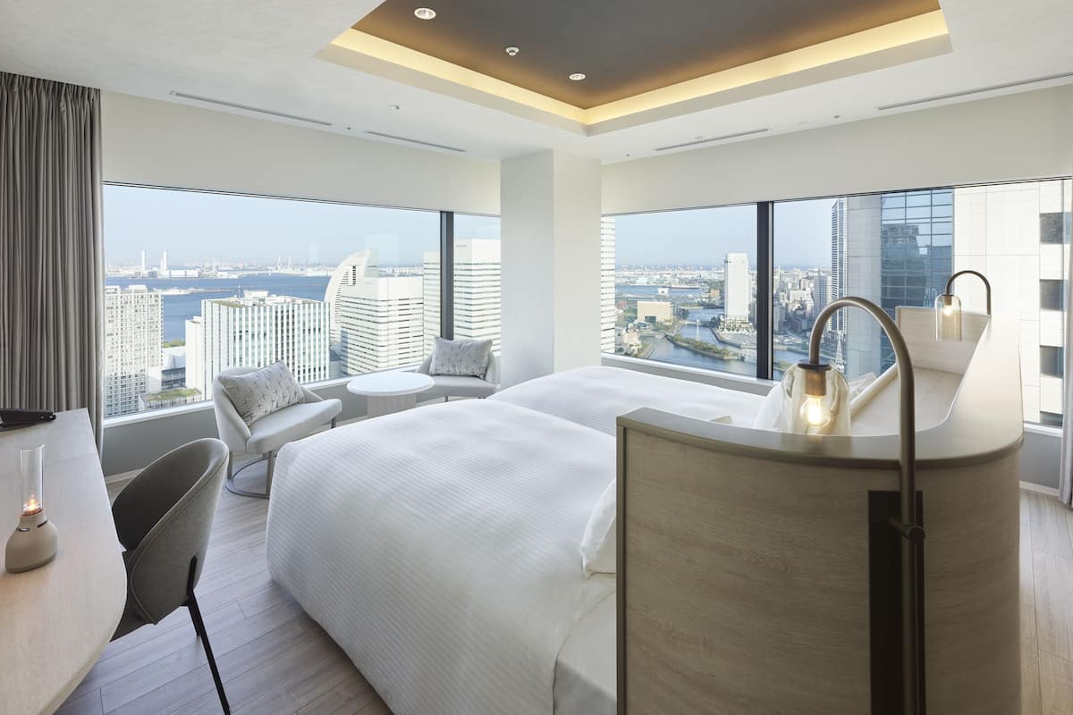 A modern hotel room with a large bed, a round table, and a seating area with two chairs. The room features a large window with a city view.