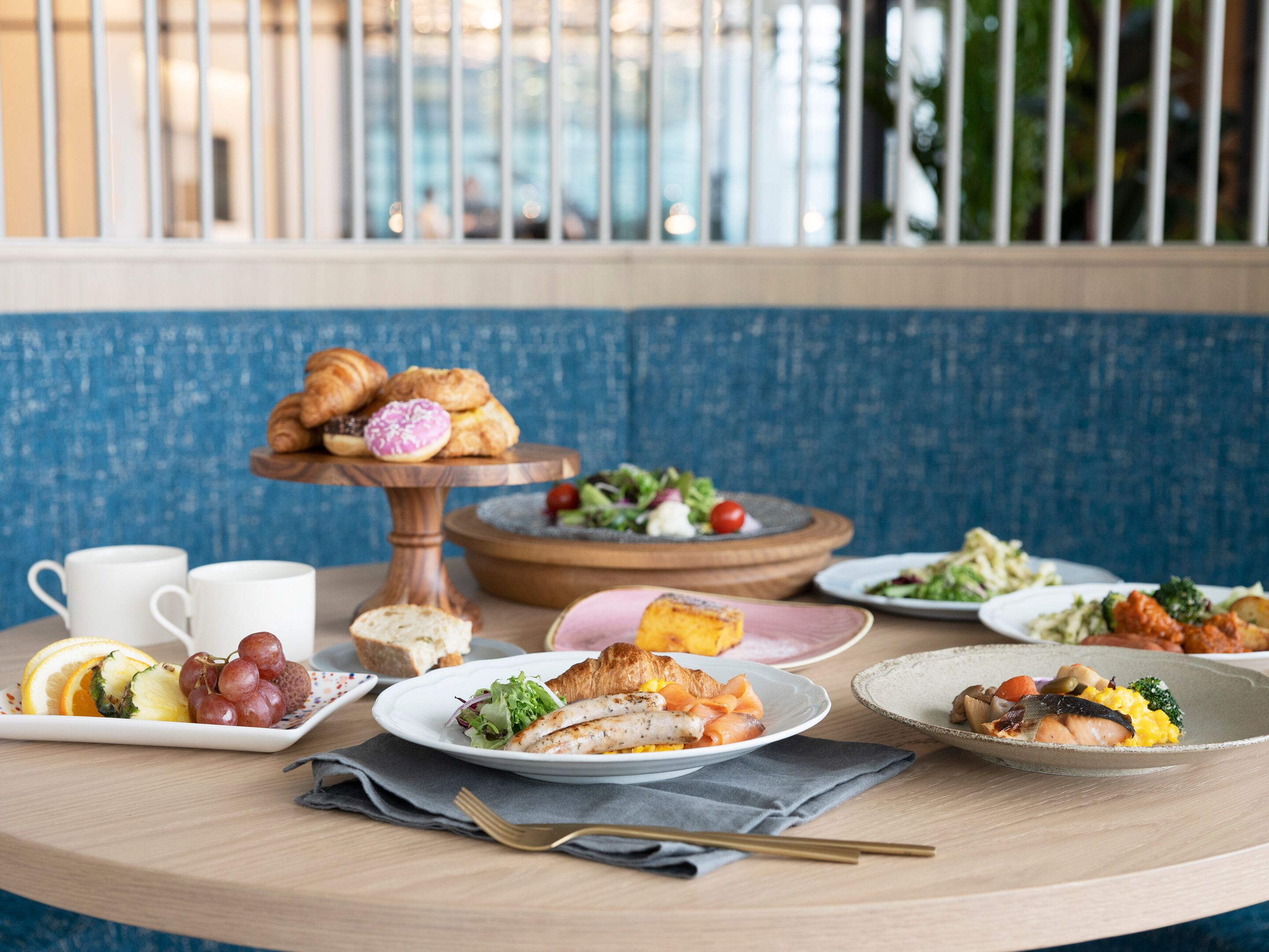 A breakfast buffet with various dishes, including croissants, fruit, and salads, on a wooden table with a blue wall in the background.
