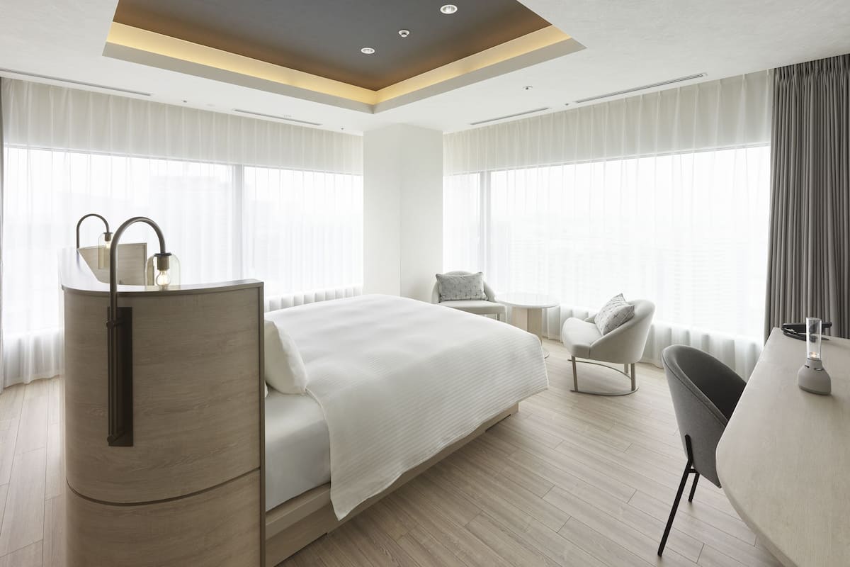 A modern hotel room with a large bed, a desk, and a chair. There are large windows with curtains, a ceiling light, and a radiator.