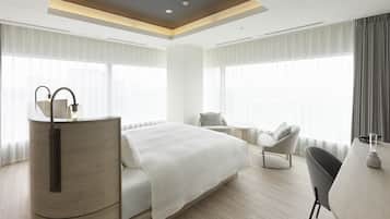 Habitación de hotel moderna con una cama grande, un escritorio y una silla. Cuenta con ventanas amplias con cortinas, una lámpara de techo y un radiador.