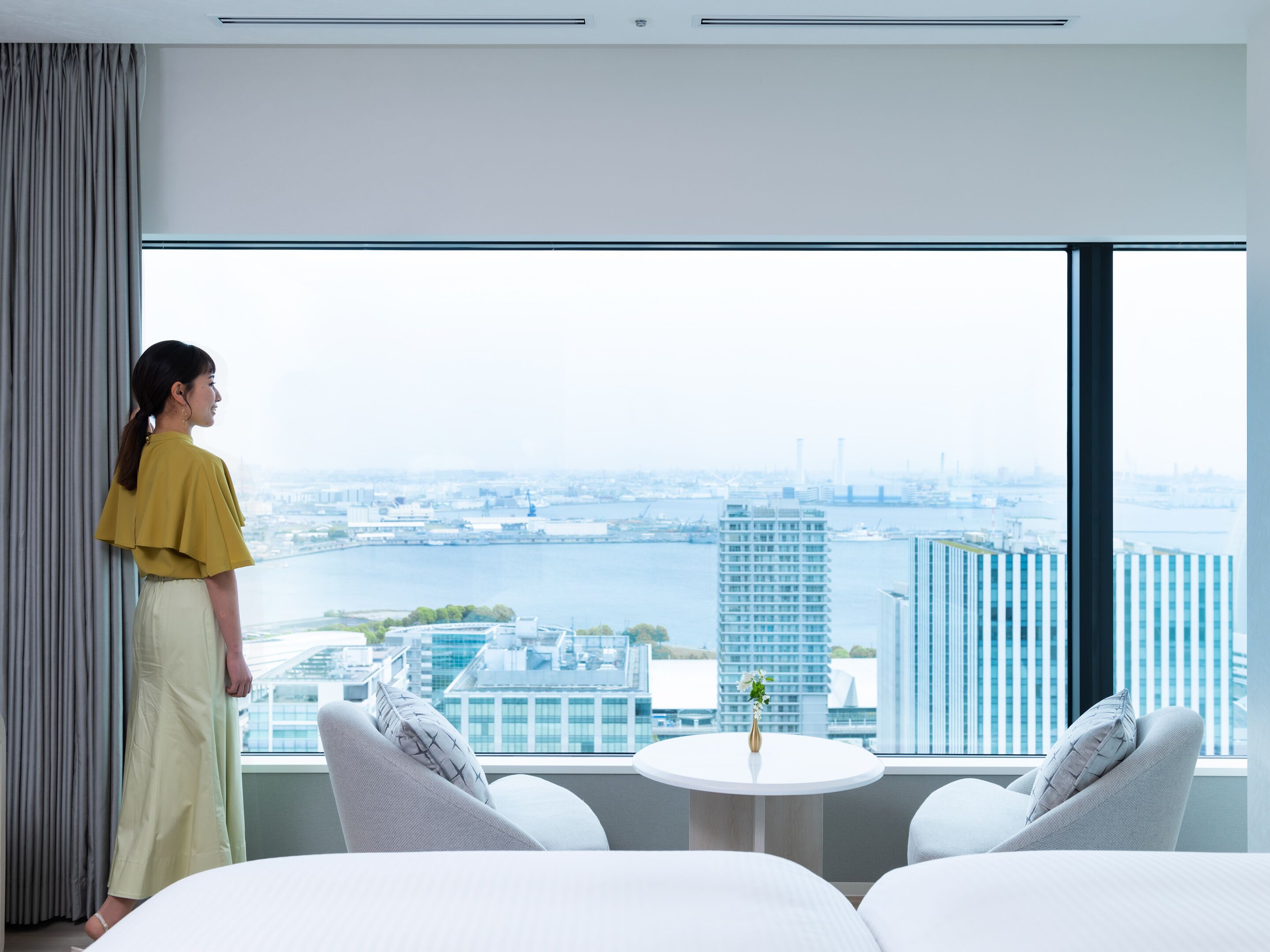 A modern hotel room with a large window offering a city view, a small round table with a vase, and two grey upholstered chairs.