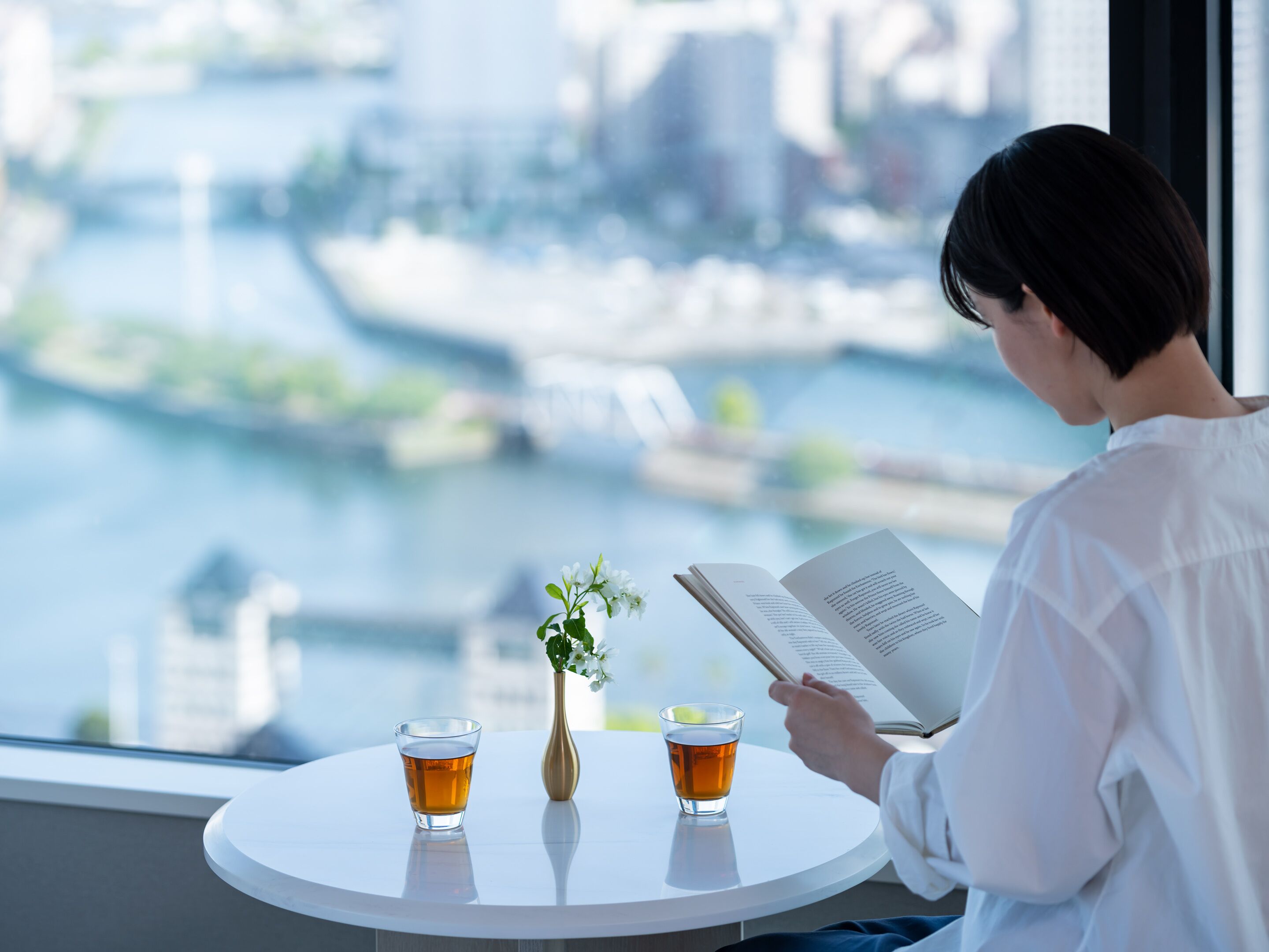 Eine Person liest ein Buch an einem Tisch, neben dem zwei Teegläser und eine Blumenvase stehen. Durch das Fenster ist ein Fluss zu sehen.