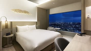 A modern hotel room with a large bed, bedside tables, a desk, and a view of a cityscape at night.