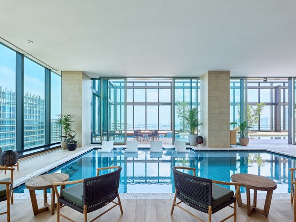 Ein moderner Poolbereich im Innenbereich mit großen Glaswänden, Holzboden und mehreren Sitzbereichen mit Stühlen und Tischen.