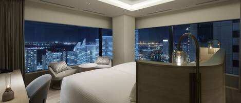 A modern hotel room with a large bed, a desk, and a view of a cityscape at night.