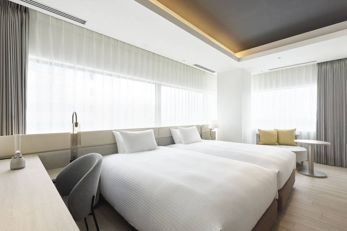 A modern hotel room with two large beds, a desk, a chair, and a small round table.