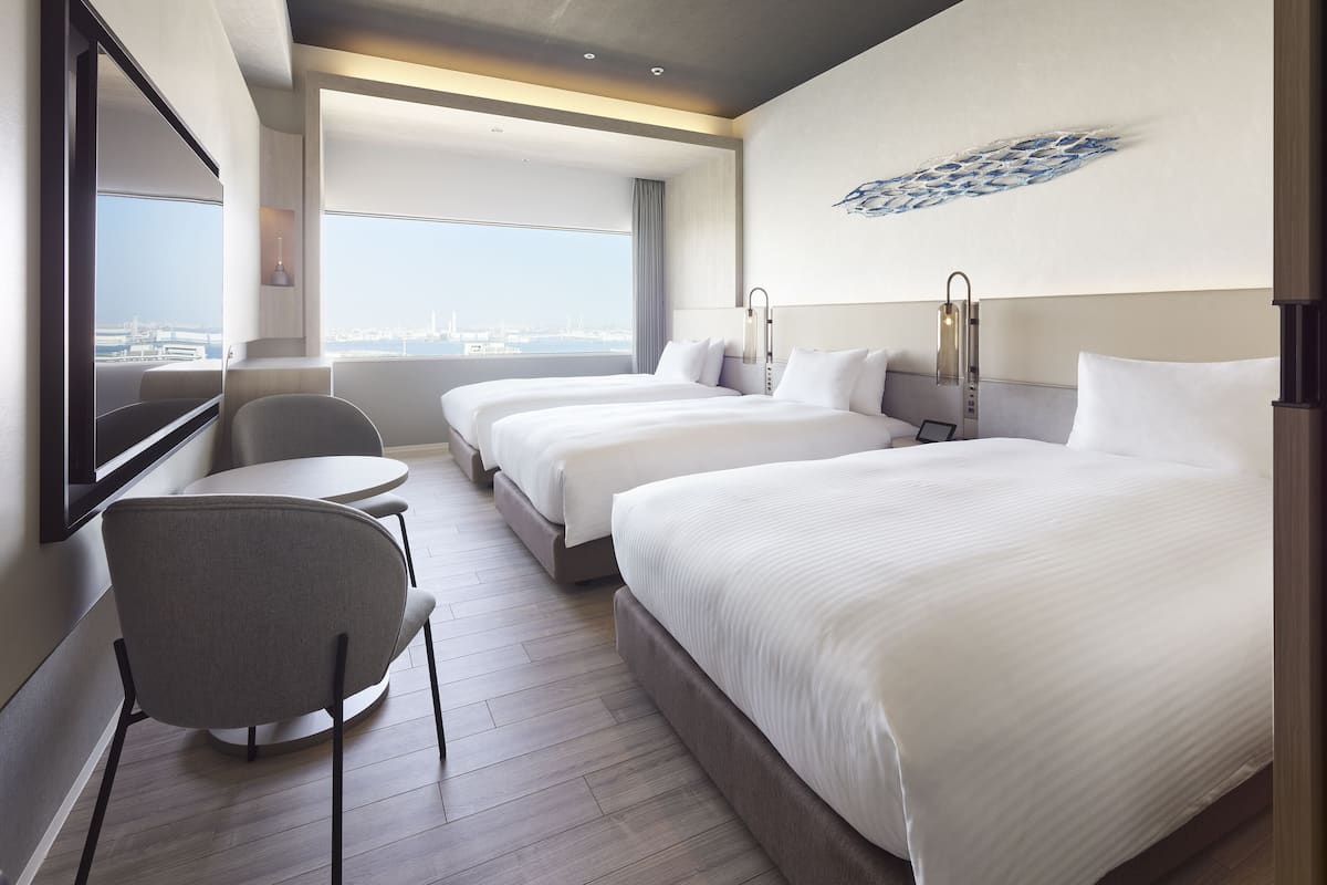 A hotel room with two beds, a desk, and a chair. There is a large window offering a view of the city.