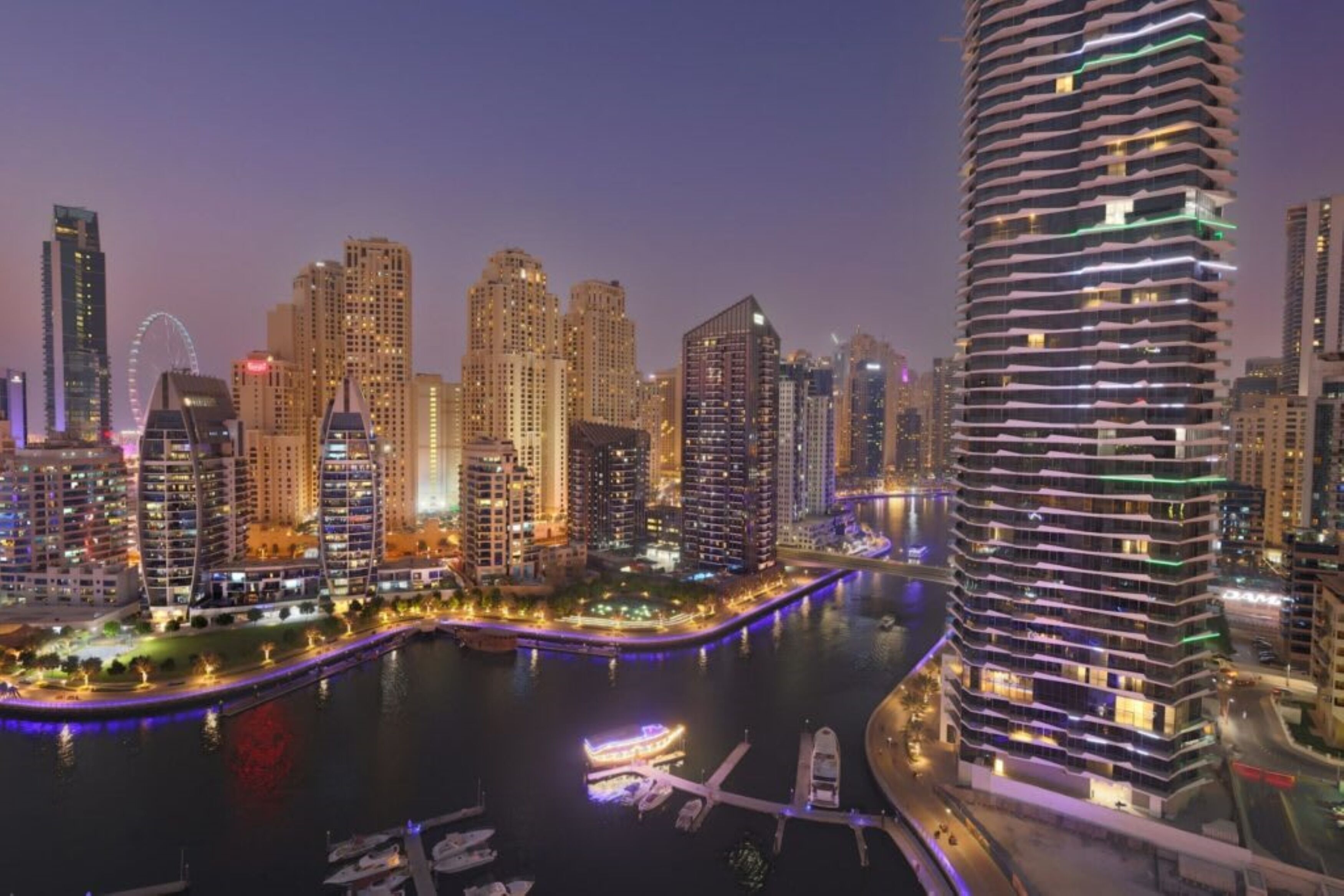 Une vue nocturne d’une ville, avec un front de mer bien visible, des immeubles de grande hauteur et un gratte-ciel au design distinctif, caractérisé par sa forme incurvée.