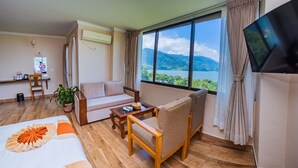 A hotel room with a sofa, armchair, wooden floor, TV, and a large window with a mountain and lake view. - Hotel Fewa Camp (Pokhara)
