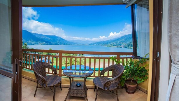 A balcony with wicker chairs and a glass table, overlooking a lake and mountains. - Hotel Fewa Camp (Pokhara)