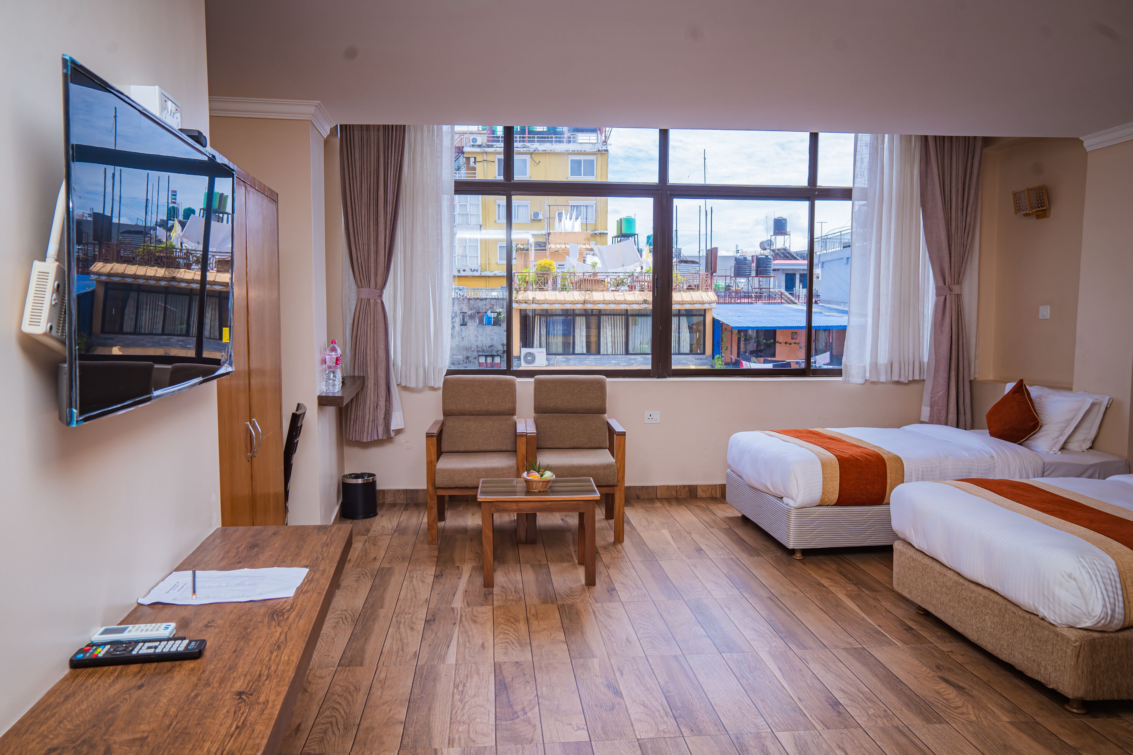 Een hotelkamer met een houten vloer, twee eenpersoonsbedden, een slaapbank, een televisie aan de muur en een raam met uitzicht op gebouwen.