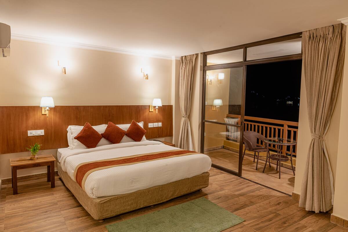 A hotel room with a large bed, two bedside lamps, a nightstand, a small table with a plant, and a balcony with a chair and table.