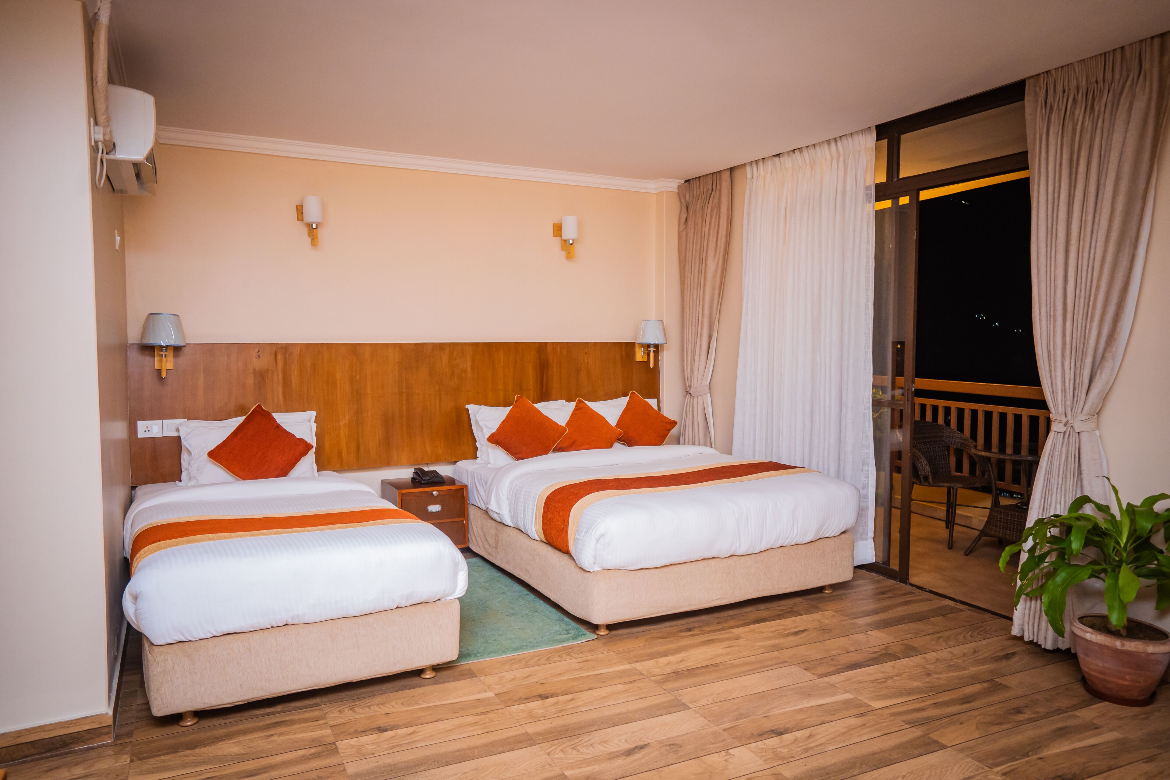 A hotel room with two beds, wooden flooring, a nightstand, a potted plant, and a balcony with curtains.
