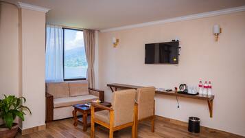 Una habitación con sofá, una mesa de madera, dos sillas, televisor montado en la pared, planta en maceta y estante con botellas y una tetera.