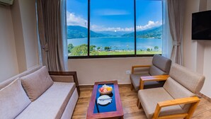 A room with a sofa, armchair, and a table with fruit. A large window offers a view of a lake and mountains. - Hotel Fewa Camp (Pokhara)