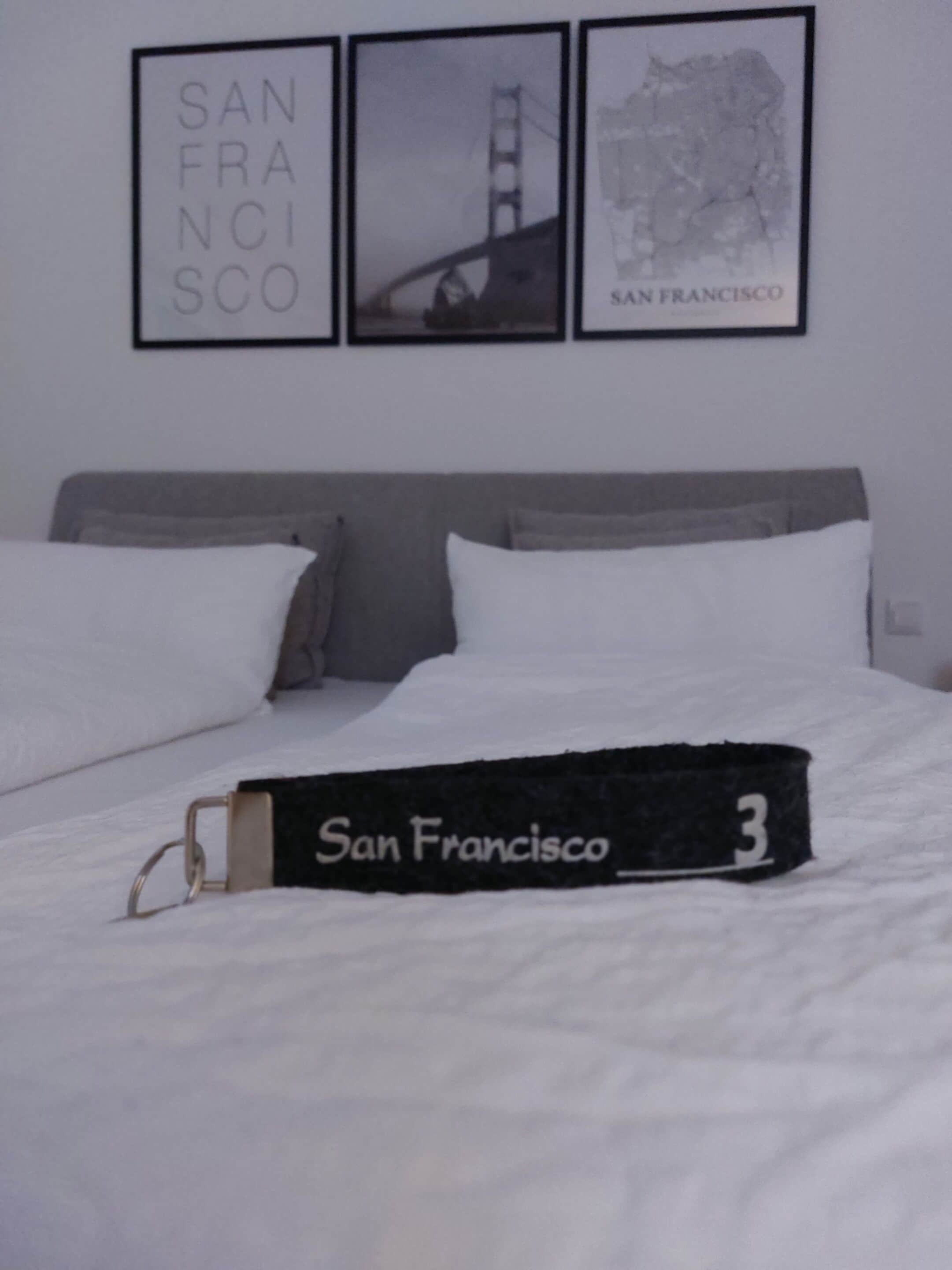 Cama com roupa de cama e travesseiros brancos, um travesseiro com tema de São Francisco e três quadros na parede acima da cama.