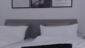 Cama com roupa de cama e travesseiros brancos, um travesseiro com tema de São Francisco e três quadros na parede acima da cama.