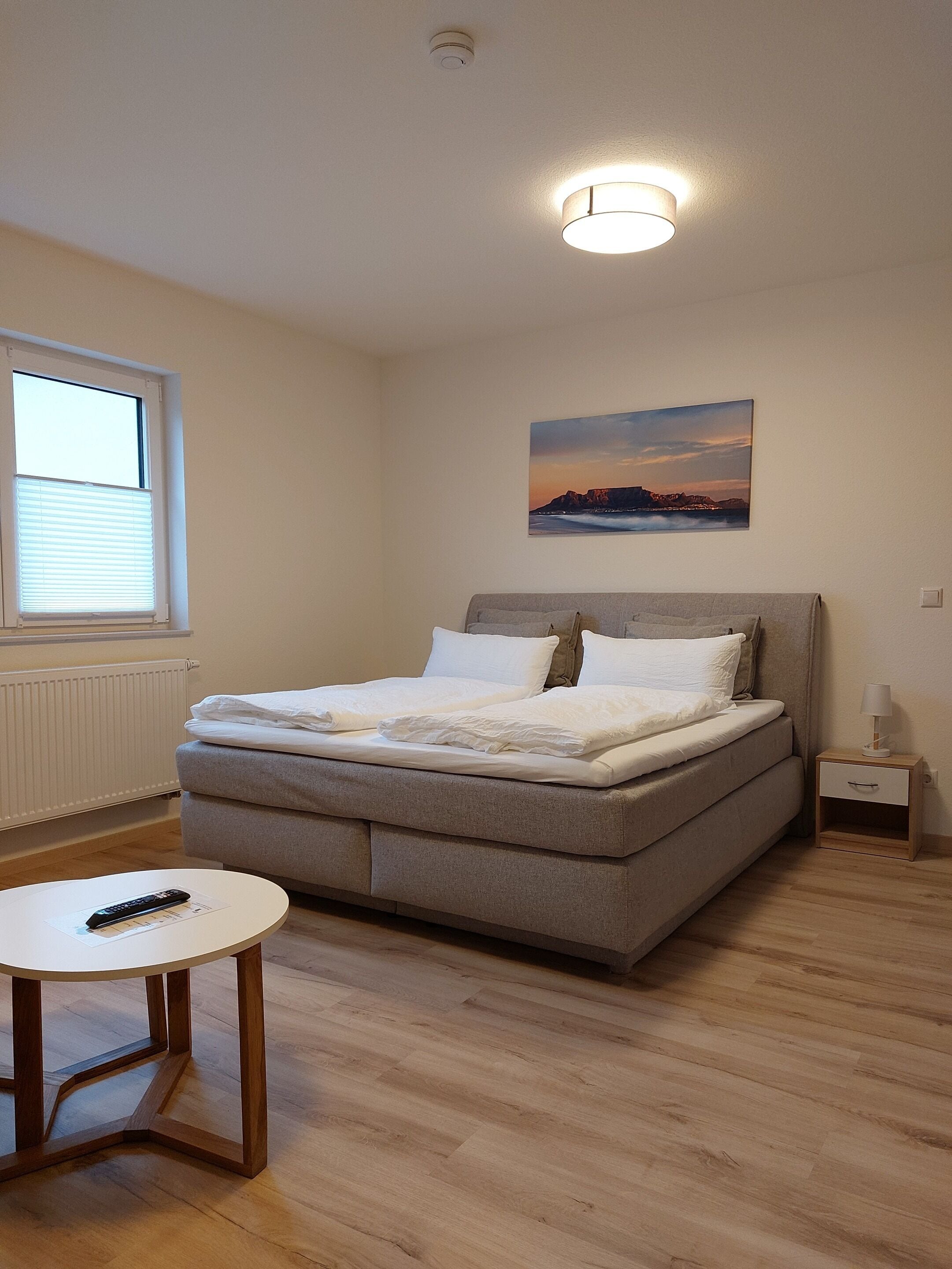 Une chambre à coucher avec un lit, une table de chevet, une lampe, une fenêtre avec des stores et un tableau au mur.