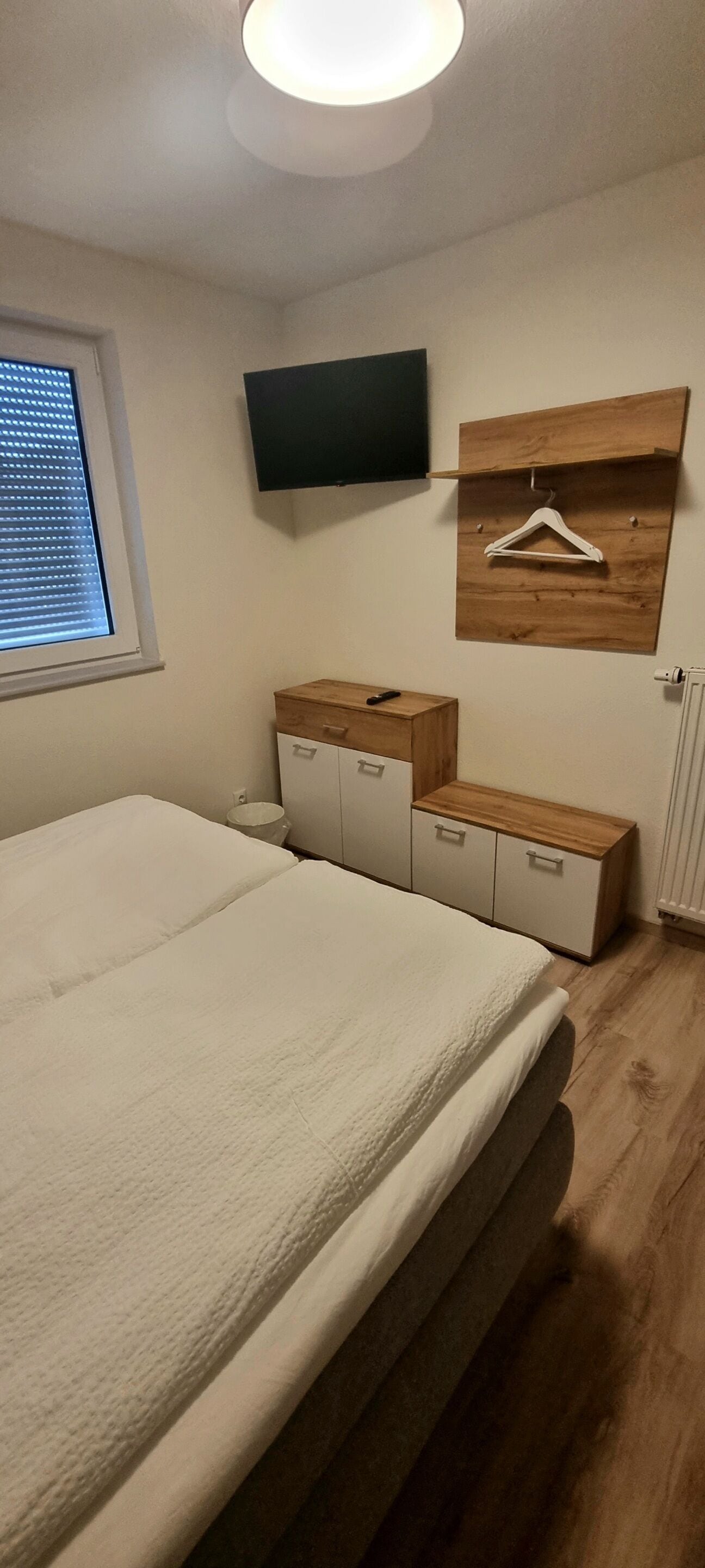 A bedroom with a large bed, a wooden dresser, a TV mounted on the wall, and a window with blinds.