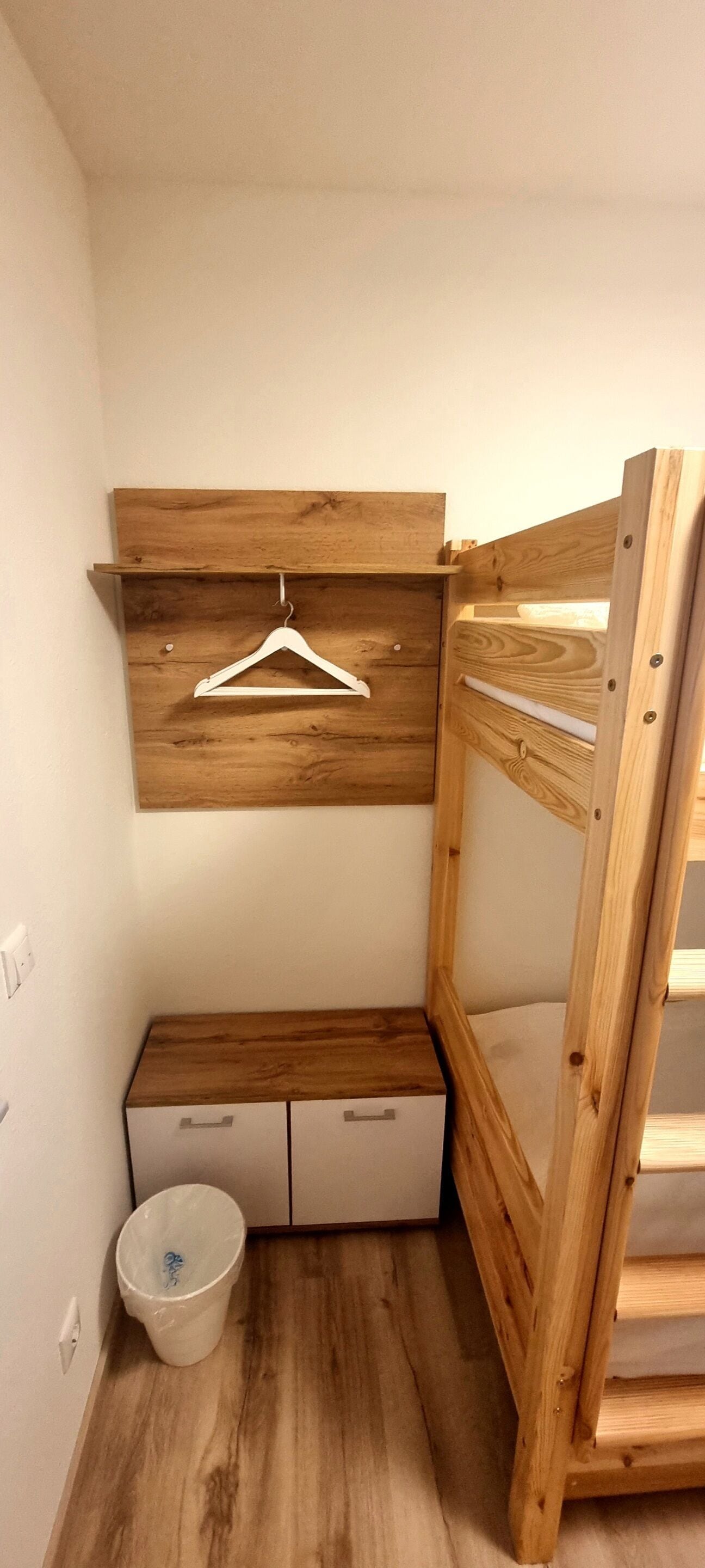 A small, compact room with a wooden bunk bed, a white dresser, a hanging clothes rack, and a trash bin.