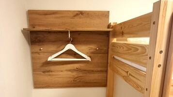 A small, compact room with a wooden bunk bed, a white dresser, a hanging clothes rack, and a trash bin.