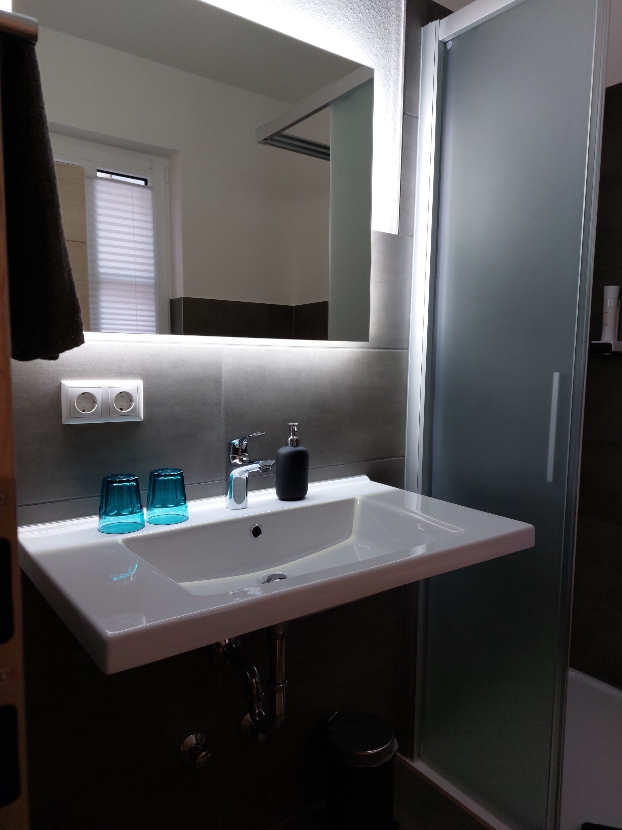 Ein Badezimmer mit einem weißen Waschbecken, zwei blauen Glasbechern, einem schwarzen Seifenspender und einem Spiegel über dem Waschbecken.