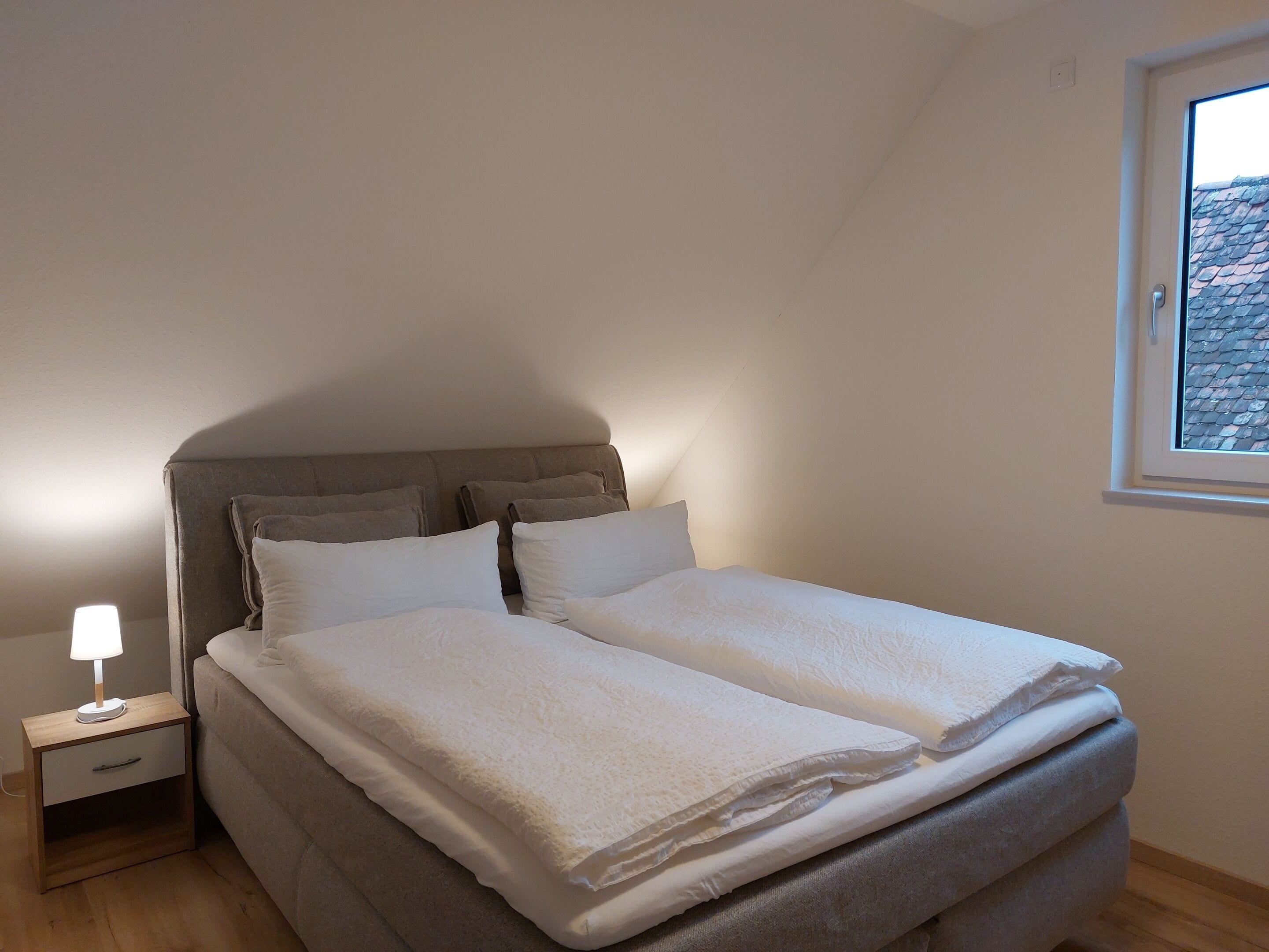 Un dormitorio con una cama, una mesita de noche con lámpara y una ventana.