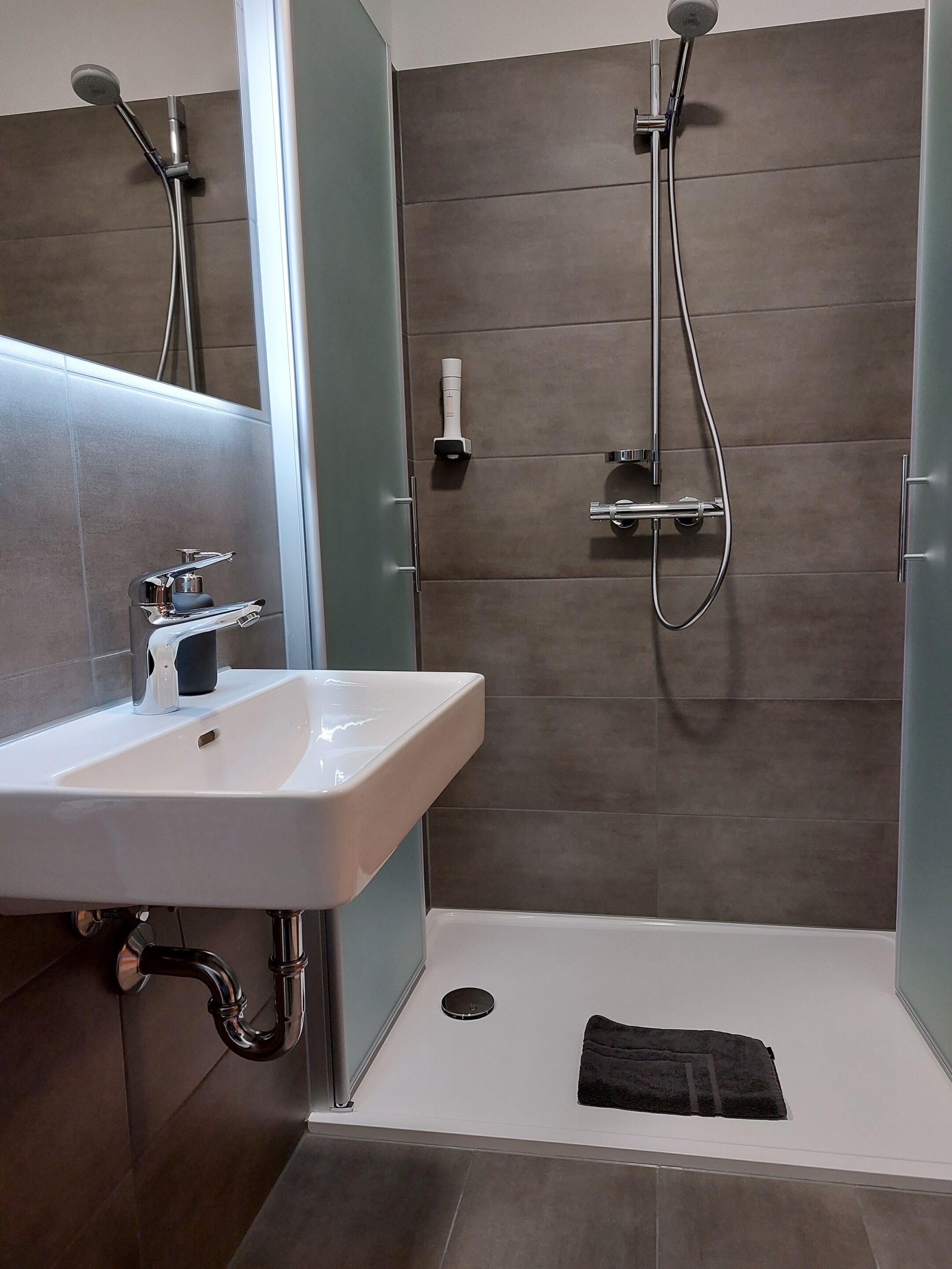 A modern bathroom with a white sink, a handheld showerhead, a built-in toilet, and a black towel on the floor.