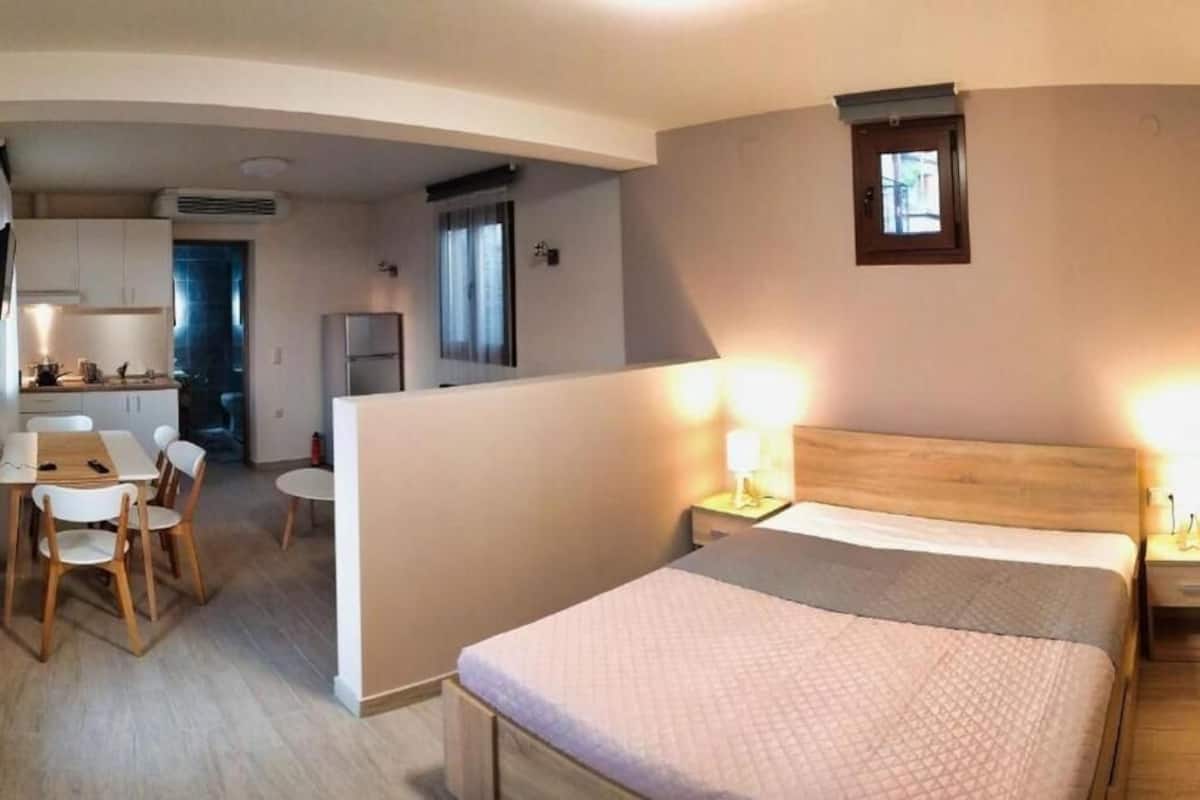 A compact hotel room with a bed, bedside lamps, a small table, and a chair. There is a kitchenette in the background with a refrigerator and a window with curtains.