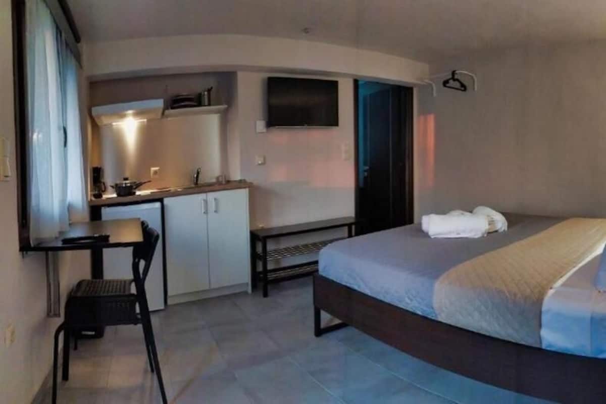 A compact hotel room with a bed, a small desk, a kitchenette, and a TV.