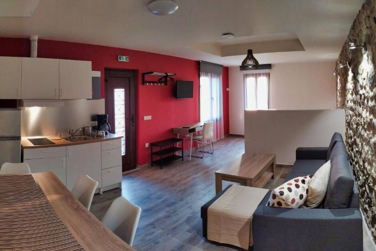 A modern kitchen with white cabinets, a dining area with a wooden table and chairs, and a living area with a sofa and a patterned cushion.