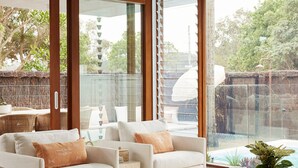 Zona relax esterna moderna con divani e cuscini bianchi, un pannello di vetro e vista su giardino e barbecue.