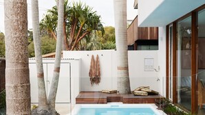 Area piscina moderna all'aperto con solarium in legno, un muro bianco con una porta e un'entrata vetrata verso un portico coperto.