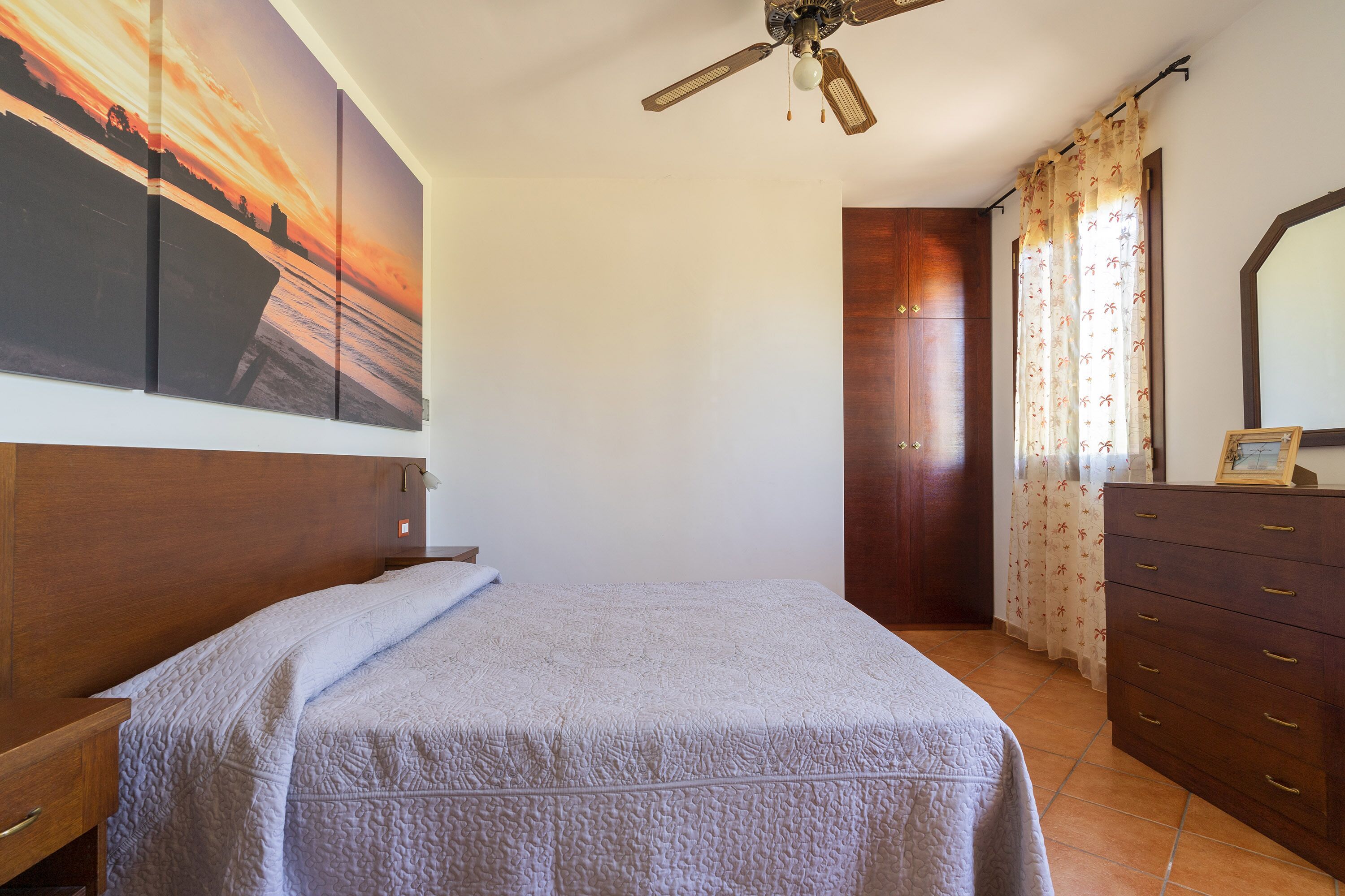 A bedroom with a bed, a ceiling fan, a wooden dresser, a large painting, and a window with curtains.