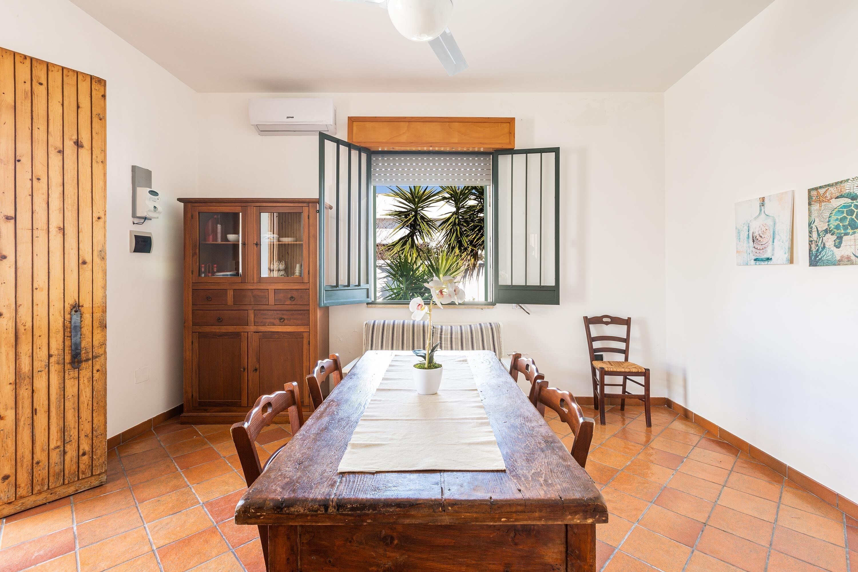A dining area with a wooden table, chairs, a cabinet, and a window with a view of palm trees.