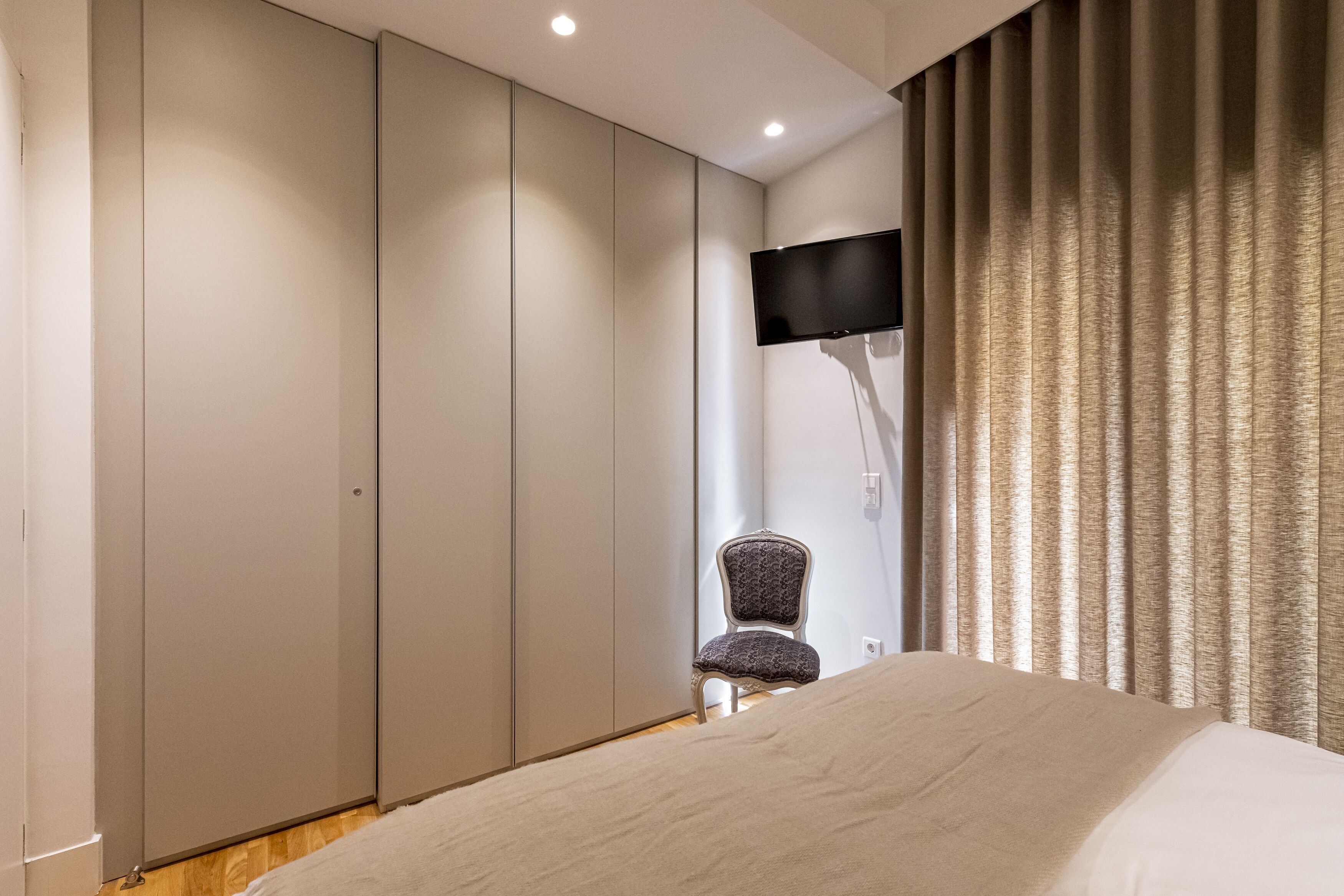 A bedroom with a bed, a chair, a TV mounted on the wall, and a curtain.