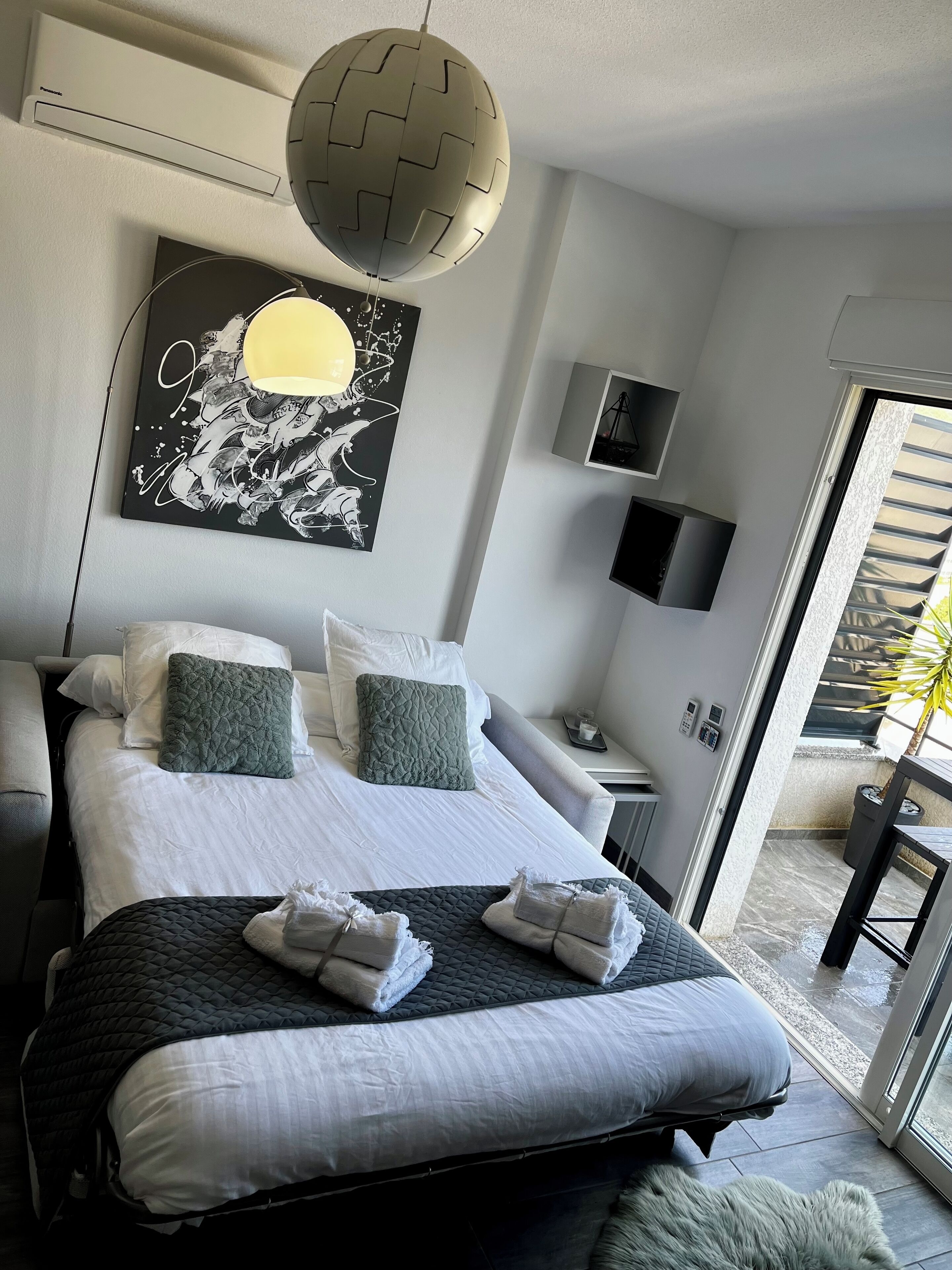 Une chambre d’hôtel moderne dotée d’un grand lit, d’un climatiseur fixé au mur, d’un luminaire sphérique original, d’une œuvre d’art encadrée, d’une petite table de chevet avec une lampe et d’une vue sur l’espace extérieur grâce à une porte coulissante.