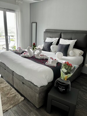 A modern hotel room with a large bed, a bedside table with a bottle, and a bouquet of flowers.