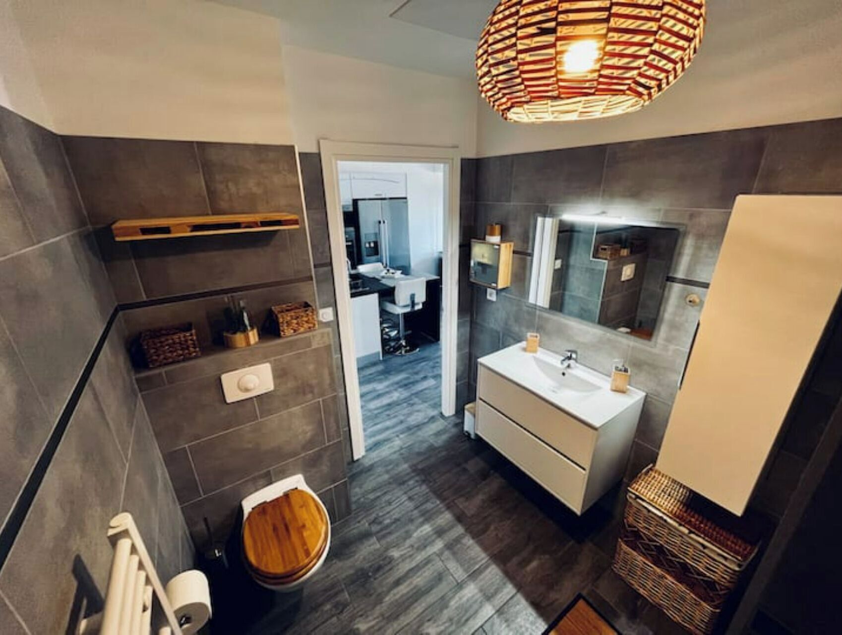 Une salle de bain moderne avec un grand miroir, un lavabo et des toilettes. On y trouve une étagère en bois avec des articles de toilette et un panier en osier.
