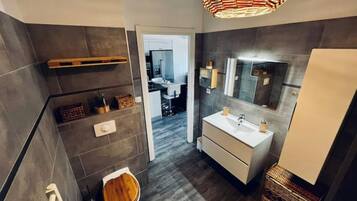 Une salle de bain moderne avec un grand miroir, un lavabo et des toilettes. On y trouve une étagère en bois avec des articles de toilette et un panier en osier.