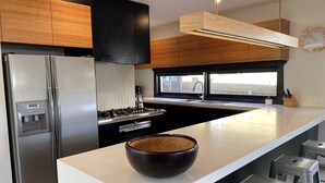 A modern kitchen with a stainless steel refrigerator, a built-in oven, a large island with a bowl, and a window providing a view. - Luxury family haven (OCEAN GROVE)