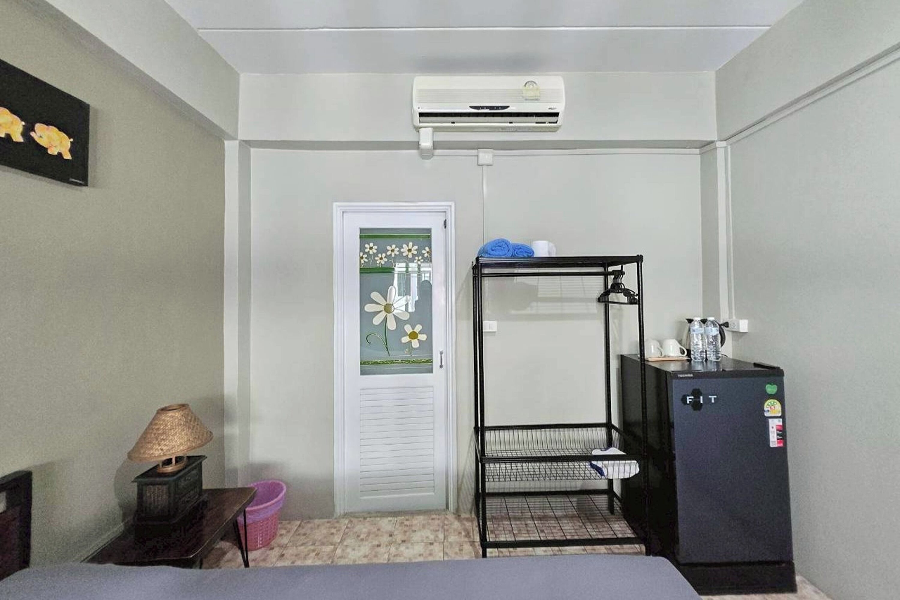 A compact room with a bed, a small table with a lamp, a black metal shelf unit, a refrigerator, and a door with a decorative glass panel.