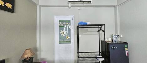 A compact room with a bed, a small table with a lamp, a black metal shelf unit, a refrigerator, and a door with a decorative glass panel.