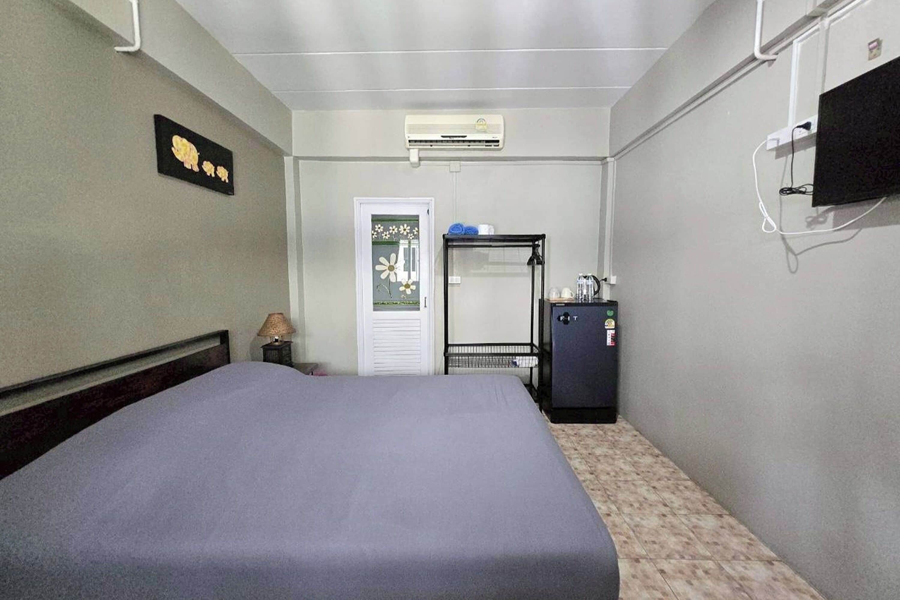 Ein Hotelzimmer mit Bett, Kleiderschrank, kleinem Tisch mit Lampe, an der Wand montiertem Fernseher und Klimaanlage.